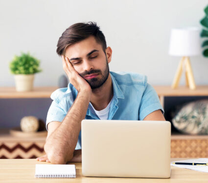 Exhausted,Millennial,Male,Hold,Head,With,Hand,Sitting,At,Office